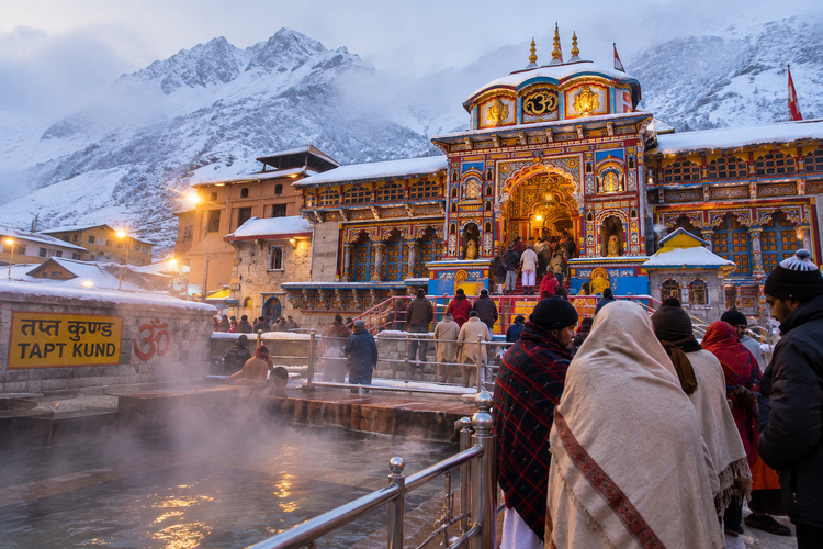 Pilgrims wrapped in warm shawls near the steaming Tapt Kund at Badrinath Temple during a cold early morning, with snow-covered Himalayan mountains in the background.